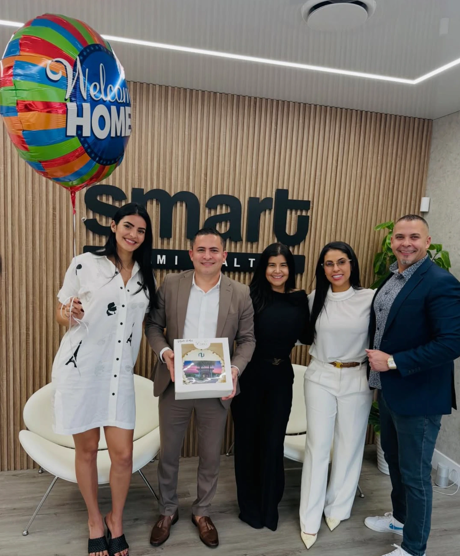 Five people celebrating a closing at the Smart Miami Realty office, beneath the Smart sign and a Welcome Home balloon.